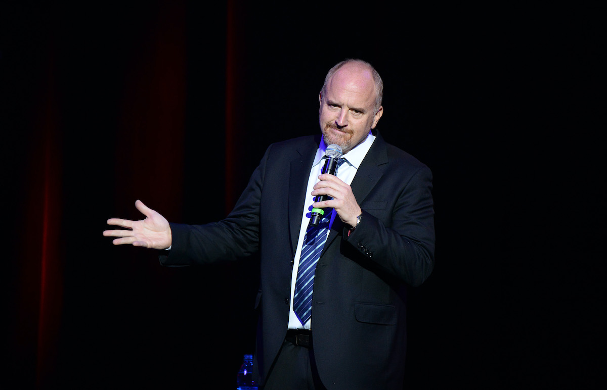 Louis CK gestures and frowns, holding a microphone during a comedy set.