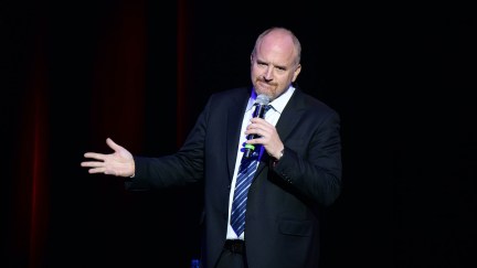 Louis CK gestures and frowns, holding a microphone during a comedy set.
