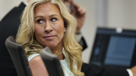 Marjorie Taylor Greene sits in a courtroom, looking over her shoulder at someone with a small smile
