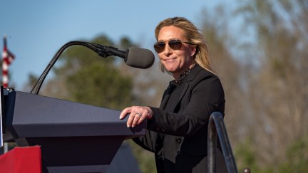 Marjorie Taylor Greene looks into the camera and smirks, wearing sunglasses, standing in front of a large podium at an outdoor rally