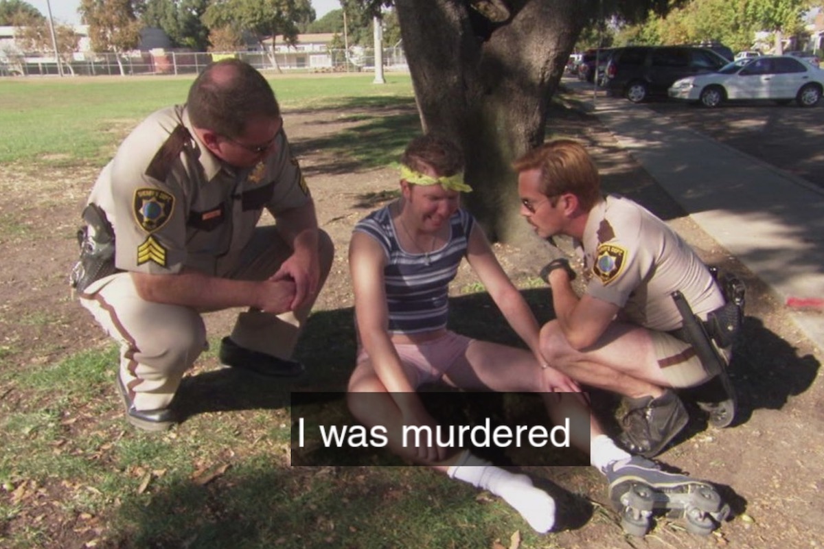 In a scene from Reno 911, a man sits on the ground between two police officers and cries, with the caption 'I was murdered'