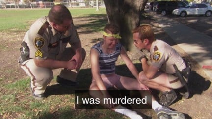In a scene from Reno 911, a man sits on the ground between two police officers and cries, with the caption 'I was murdered'