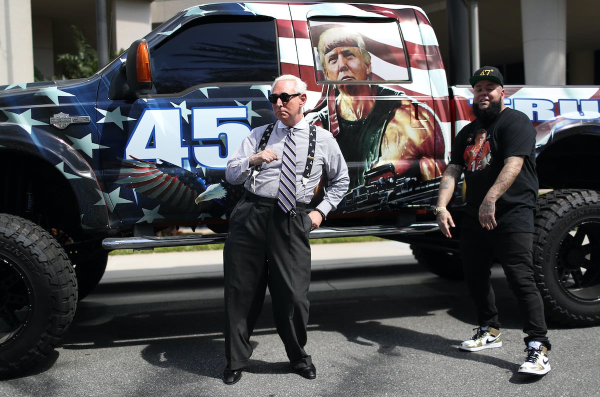 Roger Stone dances next to a rapper in front of a truck decorated with a painting of Donald Trump as Rambo