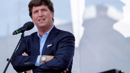 Tucker Carlson speaks into a microphone with his arms crossed, smirking