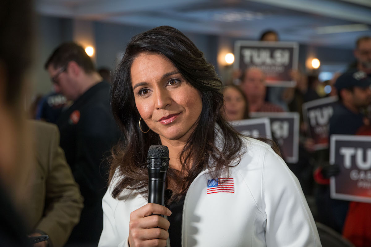 Tulsi Gabbard tilts her head and smirks, holding a microphone and speaking with a reporter