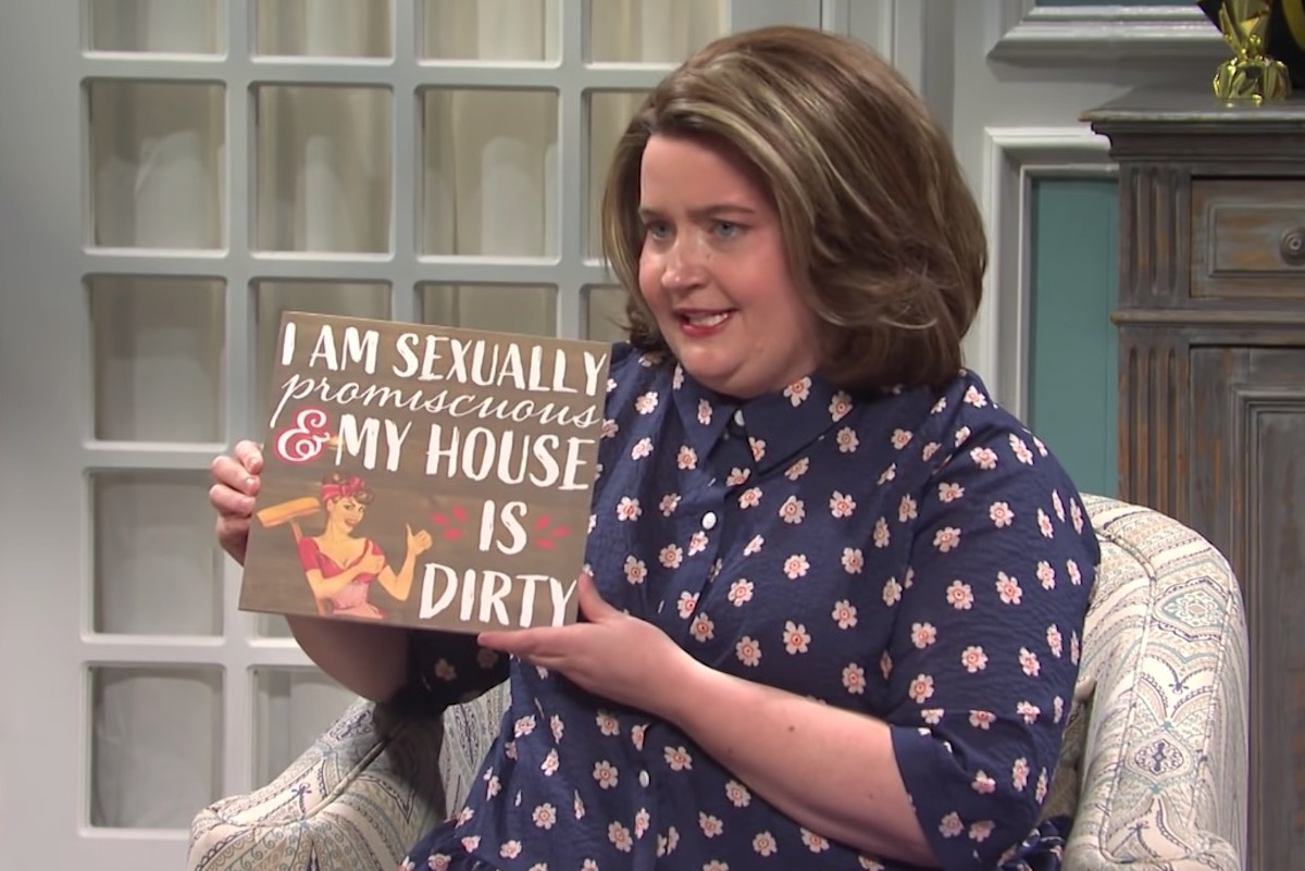 In an SNL sketch, Aidy Bryant holds a wooden sign reading "I am sexually promiscuous and my house is dirty."