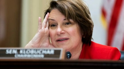 Amy Klobuchar sits looking anguished, holding her hand to her temple during a senate panel