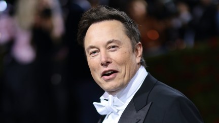 Elon Musk wears a tuxedo with a white bowtie and smiles at the camera with an open mouth