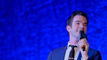 John Mulaney speaks into a microphone, standing against a blue brackdrop