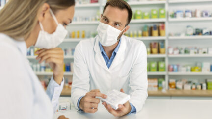 A white male pharmacist wearing a face mask talks to a white woman in a face mask about a medication