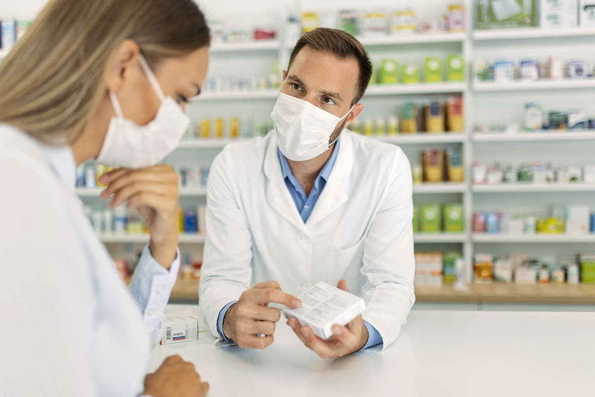 A white male pharmacist wearing a face mask talks to a white woman in a face mask about a medication