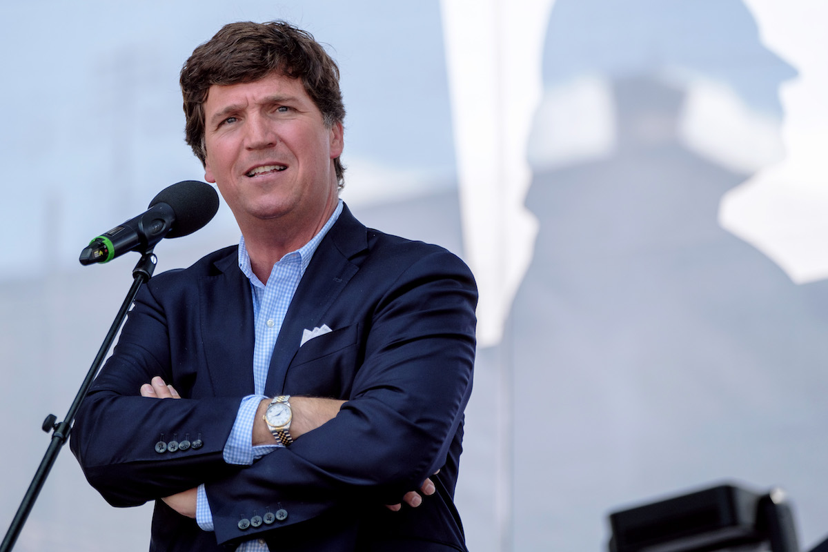 Tucker Carlson stands on a stage outside with his arms crossed, speaking into a microphone.