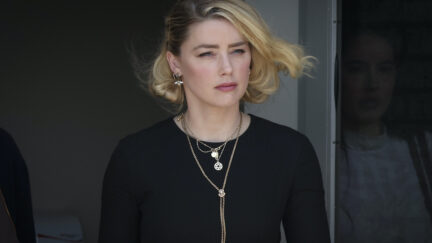 Amber Heard exits a building and the wind blows her hair, wearing a black shirt and multiple necklaces.