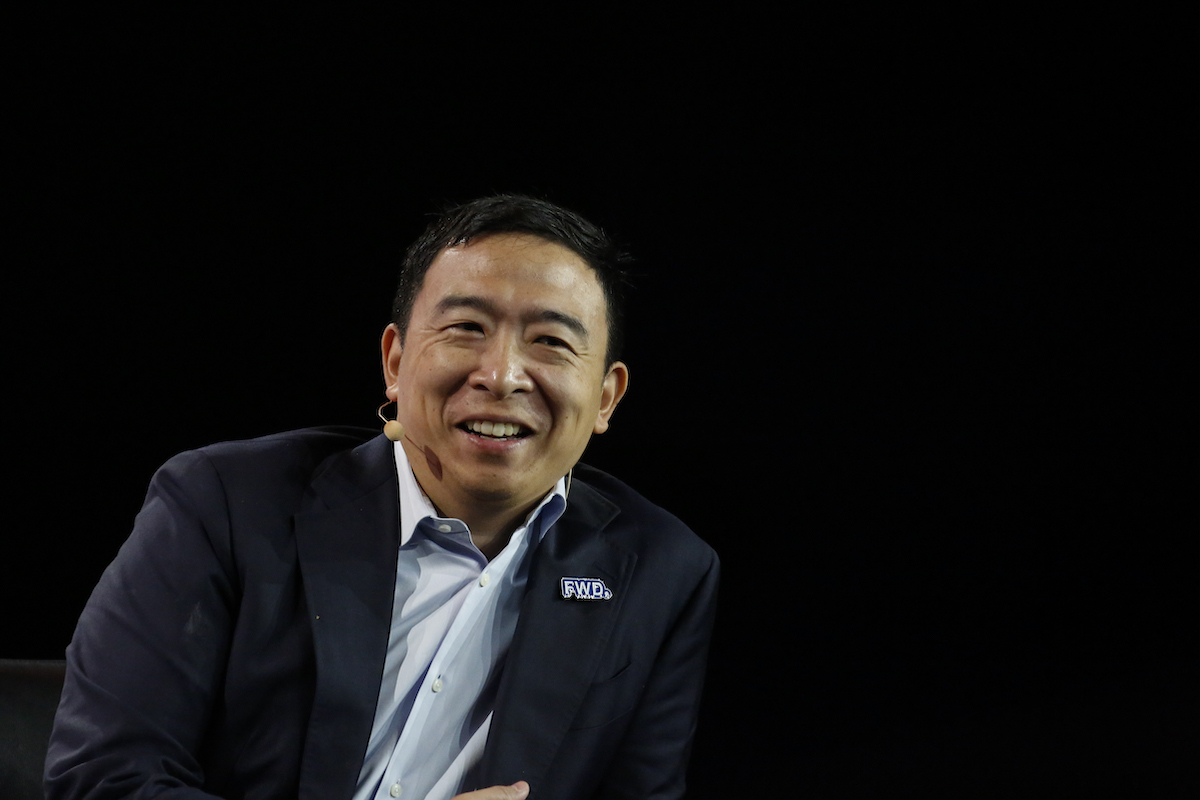 Andrew Yang speaks into a wireless microphone, sitting against a dark background