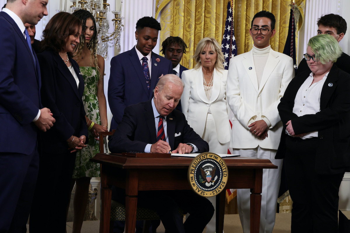Joe Biden sits at a desk signing an executive order, surrounded by LGBTQIA advocates.