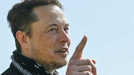 Elon Musk wears a handkerchief around his neck, holding up a finger