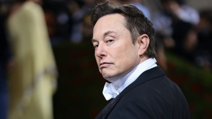 Elon Musk looks over the shoulder at the camera with a smug sort of smile, wearing a tuxedo at the Met Gala