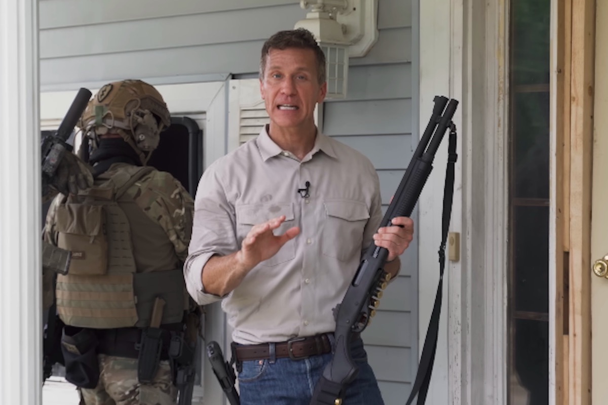 Eric Greitens holds a shotgun, gesturing and speaking into the camera, with a person in military tactical gear behind him