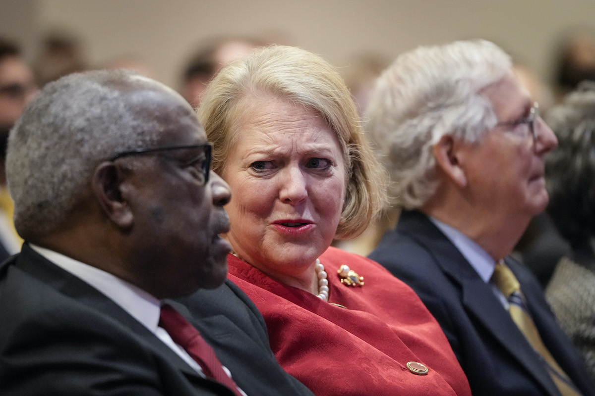 Ginni Thomas sits next to her husband Clarence THomas in the audience of an event. As the two talk to each other, she looks at him with a disbelieving expression.