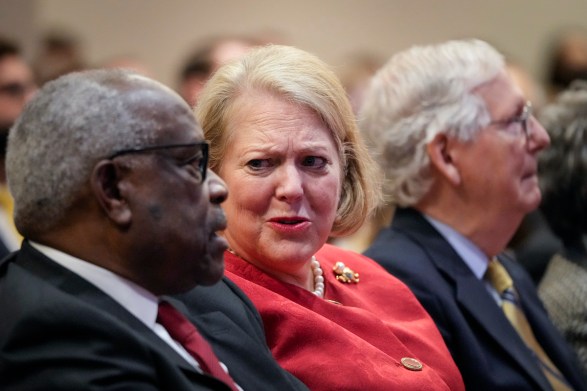 Ginni Thomas sits next to her husband Clarence THomas in the audience of an event. As the two talk to each other, she looks at him with a disbelieving expression.