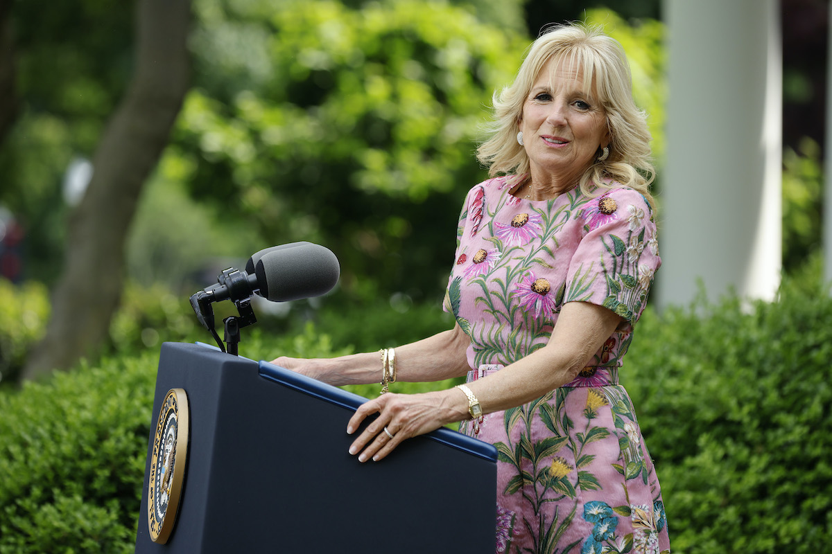 Jill Biden wears a floral dress standing at a podium, looking at the camera to her side