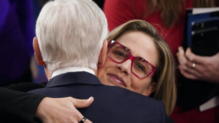 A closeup of Kyrsten Sinema's face, looking directly into the camera with a small smile as she hugs a white man with white hair.