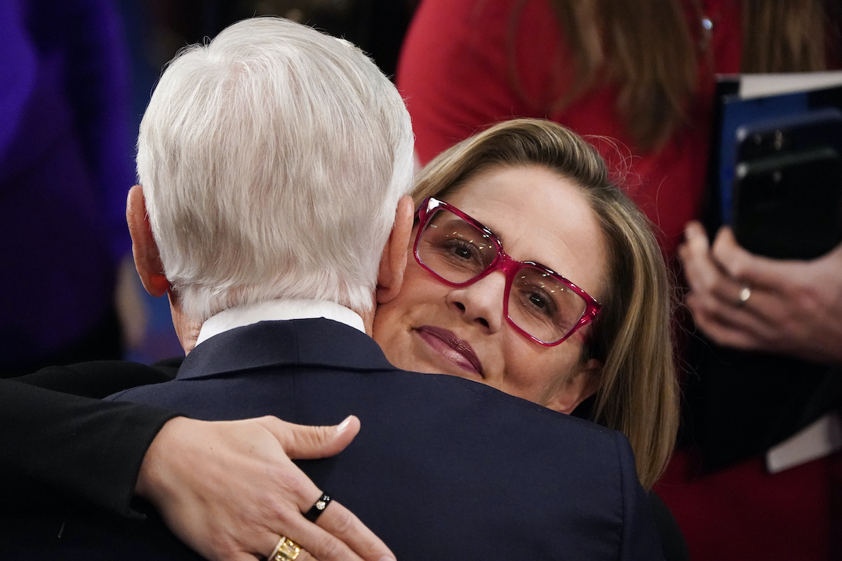 A closeup of Kyrsten Sinema's face, looking directly into the camera with a small smile as she hugs a white man with white hair.