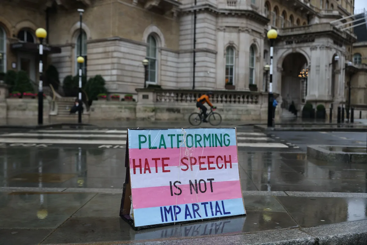 A sign colored like the blue and pink transgender flag is placed outside on a city sidewalk reading "platforming hate speech is not impartial"