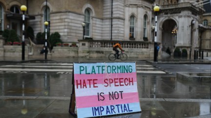 A sign colored like the blue and pink transgender flag is placed outside on a city sidewalk reading 