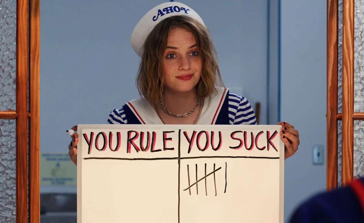 Maya Hawke as Robin in Stranger Things holding up a sign showing how much someone sucks, and it's way more than they rule.