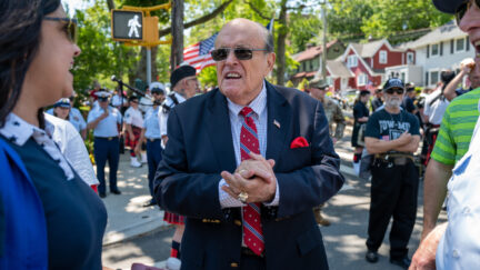 Rudy Giuliani clasps his hands and squints through sunglasses during a parade or outdoor gathering.