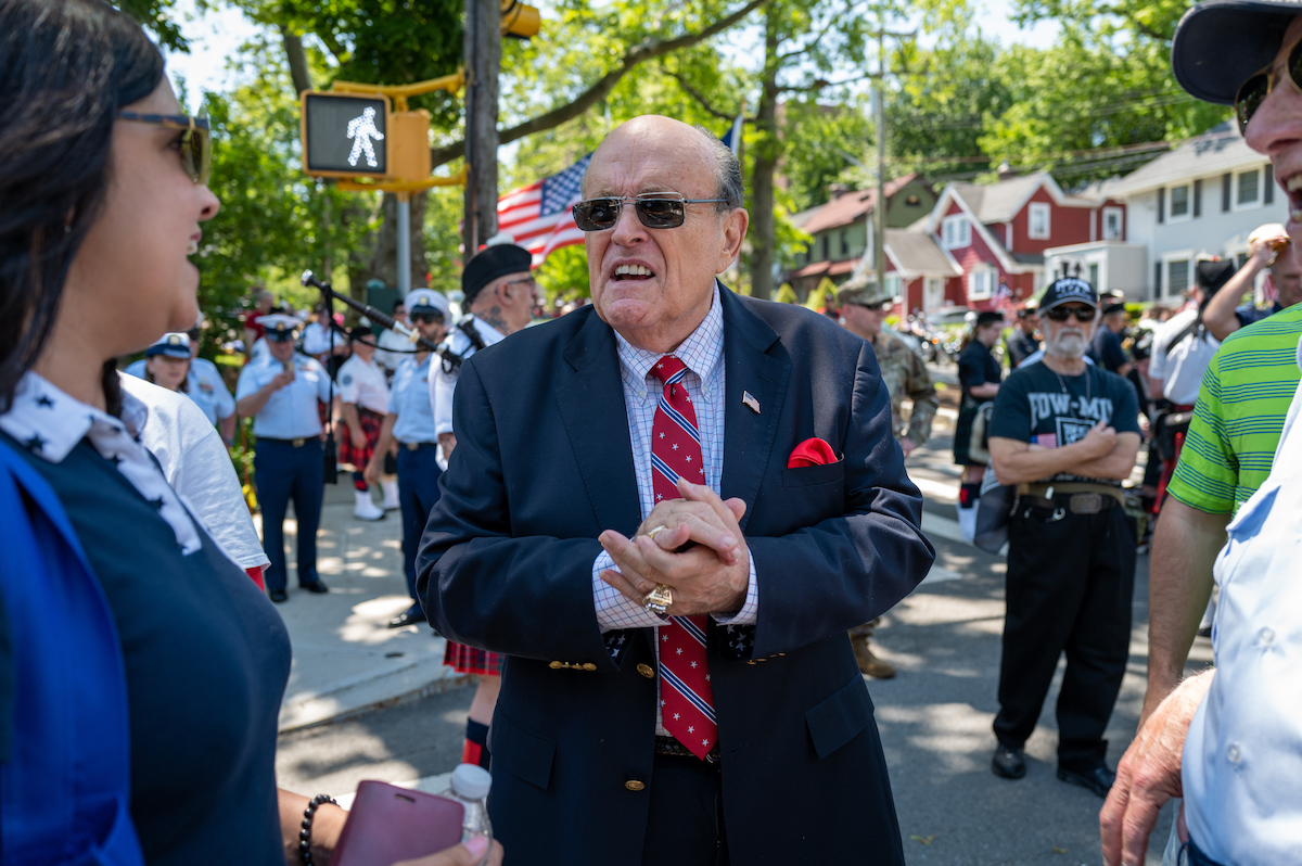 Rudy Giuliani clasps his hands and squints through sunglasses during a parade or outdoor gathering.