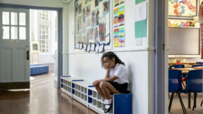A wide-angle view of a young girl with curly brown hair sitting outside of her classroom with her hand on her knee looking fed up.