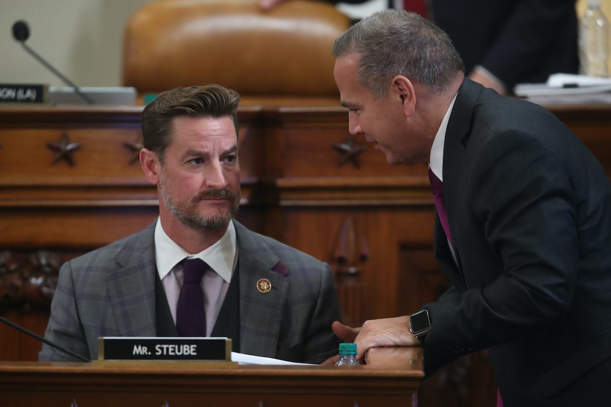 One white male US Representative leans over the desk of another white male US Rep (Steube) to talk to something. Steube listens with a blank expression.