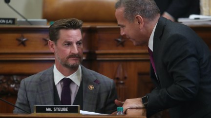 One white male US Representative leans over the desk of another white male US Rep (Steube) to talk to something. Steube listens with a blank expression.