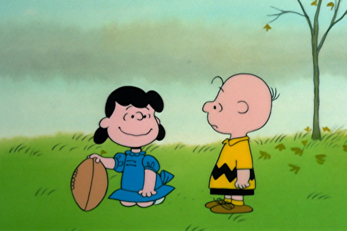 From a Peanuts cartoon, Lucy, smiling, holds a football out for Charlie Brown to kick