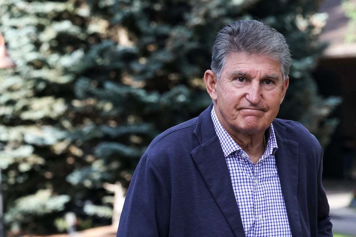 Joe Manchin has an uncomfortable small smile, standing outside in front of trees.