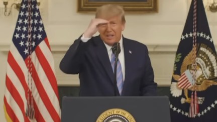 Donald Trump squints with his hand shielding his face while reading from a teleprompter from a podium