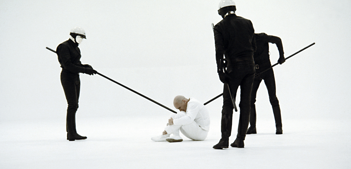 Film still from THX 1138 featuring Robert Duvall as THX 1138 crouched on the floor surrounded by android police using lances to prevent him from moving