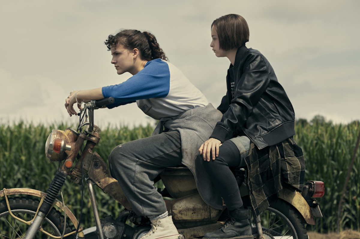 KJ, played by Fina Strazza, and Mac, played by Sofia Rosinsky, riding a motorcycle together in the countryside