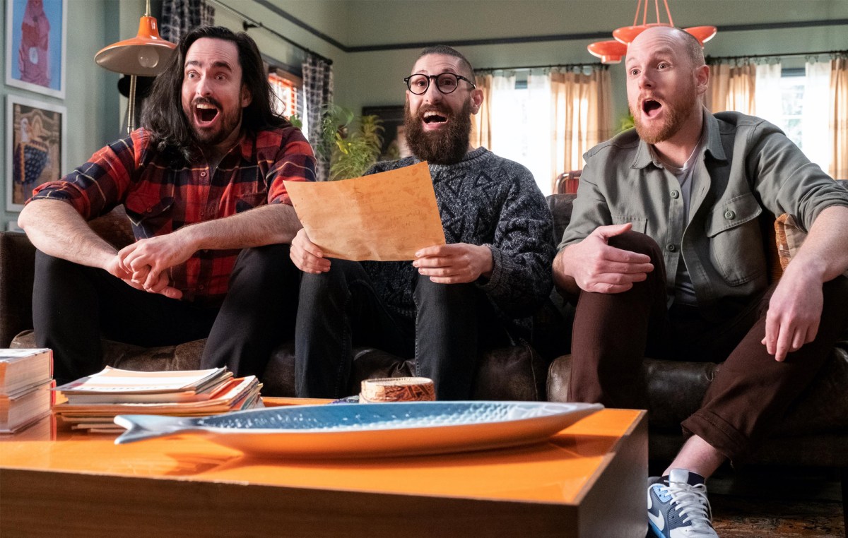 Zach, Mark, and Broden sit on their couch smiling in 'Aunty Donna's Big Ol' House of Fun.' Mark holds a brown piece of paper.
