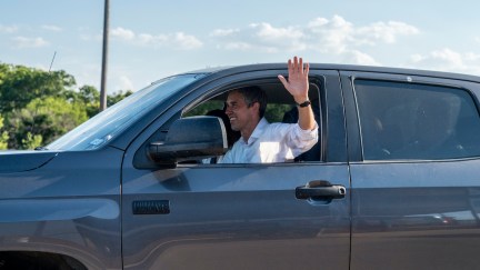 Beto O'Rourke smiles and waves out the window as he drives away in a large truck.