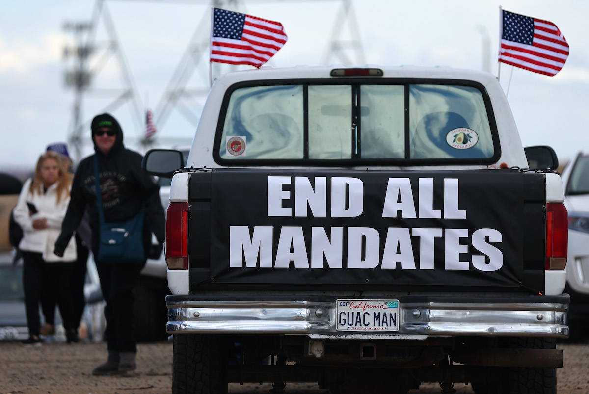 The back of a pickup truck has "end all mandates" written on it in big block letters. White men and an American flag are also in frame.