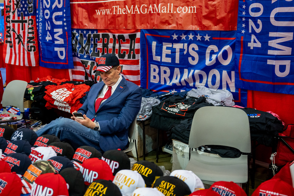 An older white man sits among rows of MAGA merchandise at CPAC.