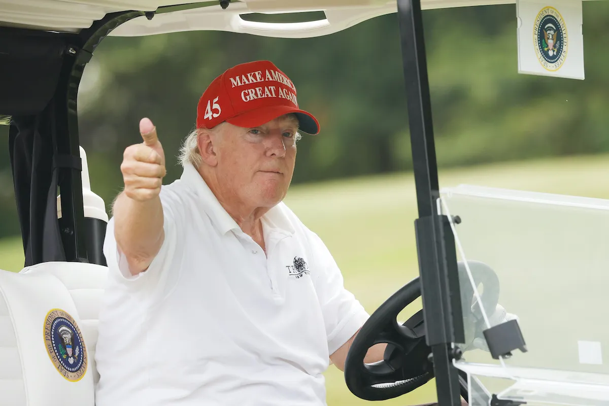 Donald Trump wears a red MAGA hat while driving a golf cart, gives a thumbs up to the camera