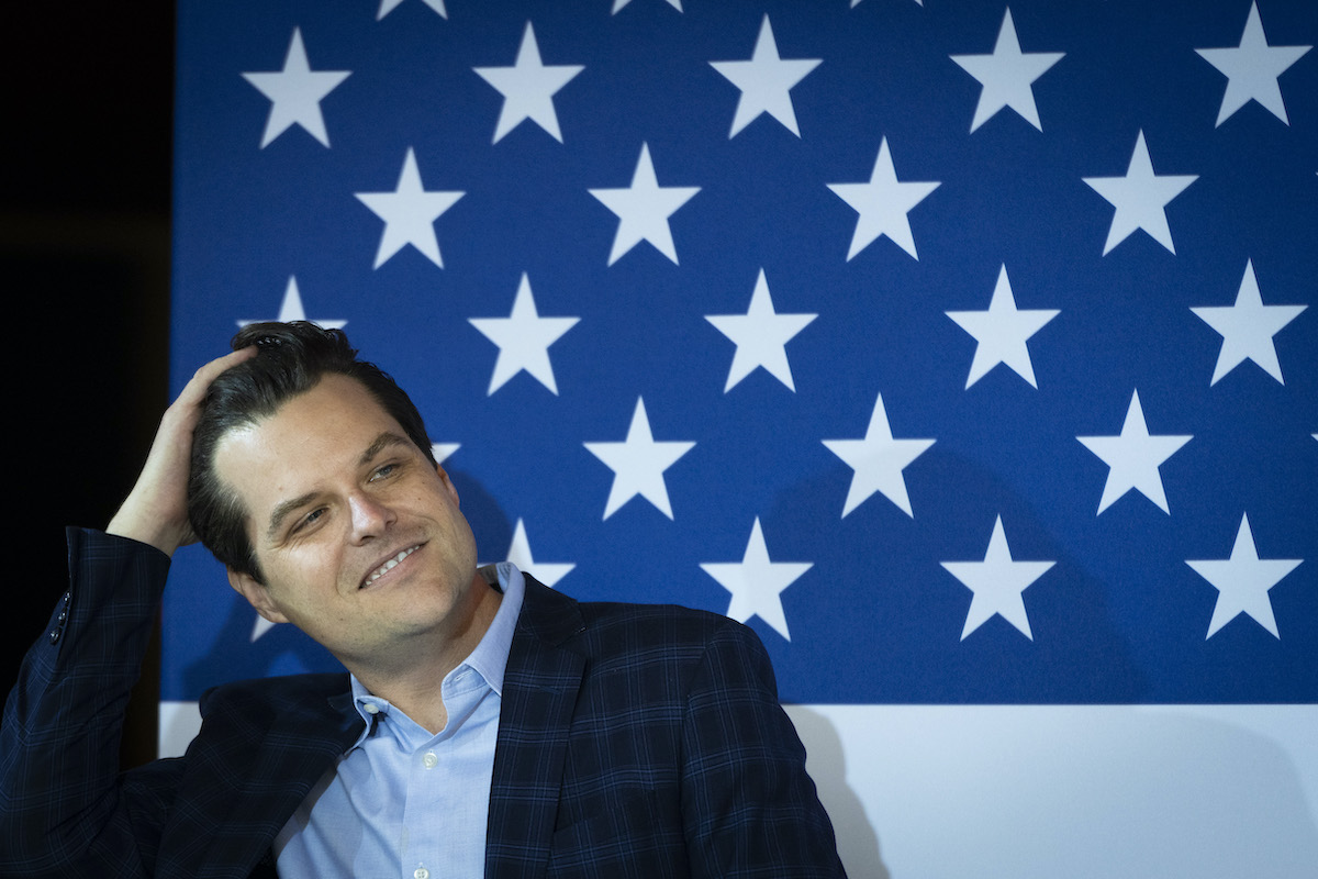 Matt Gaetz runs his hand through his hair and gives a sleazy smile sitting in front of a giant American flag