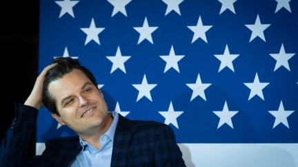 Matt Gaetz runs his hand through his hair and gives a sleazy smile sitting in front of a giant American flag