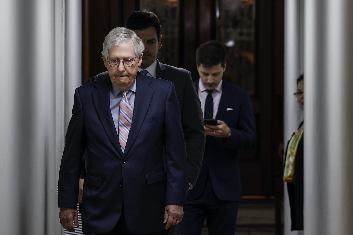 Mitch McConnell bites his lip and looks down as he walks with aides behind him.