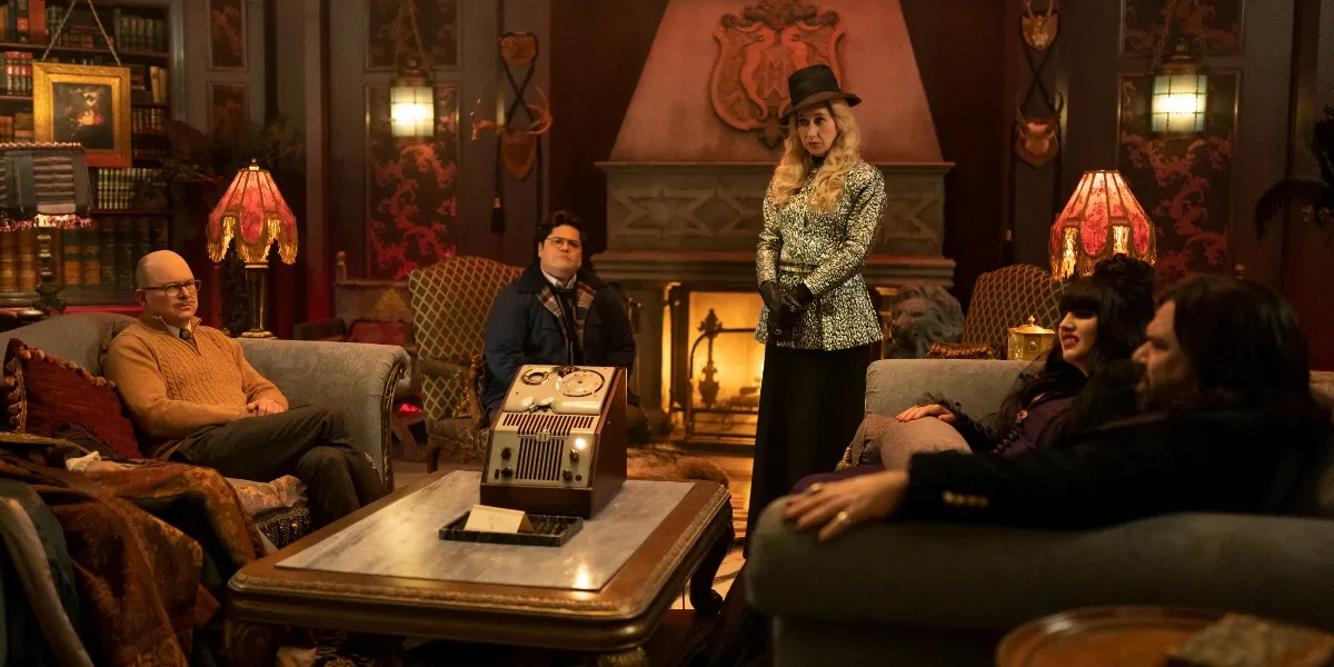 The cast of What We Do in the Shadows sits in their living room, with the Guide standing in the center.