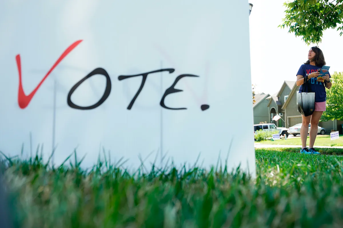 A close-up of a sign on a lawn reading "Vote," with a woman in the background